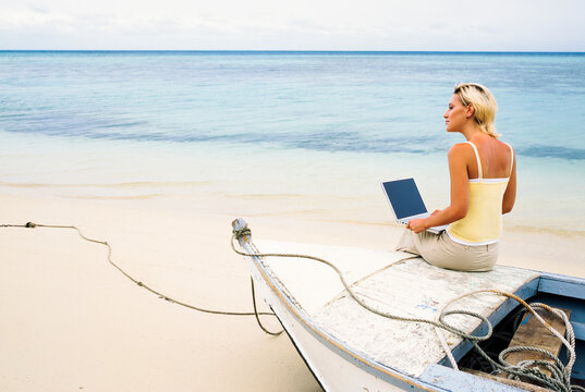 Young woman sitting on the bow of a small boat with her laptop computer on a tropical beach