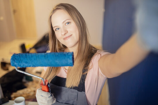 Painter happy young woman making selfie photo and painting wall with blue color paint roller in house