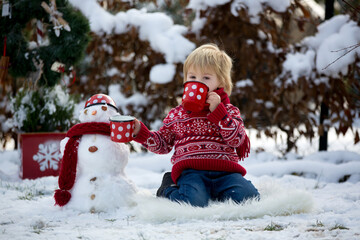 Sweet blond toddler child, boy, playing in garden with snow, making snowman, happy kid winter time