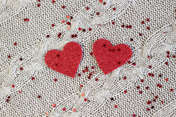Two shiny hearts cut out of red paper on a cozy knitted background with red sparkles