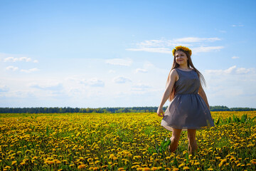 Fototapeta premium Beautiful young woman on a field with green grass and yellow dandelion flowers in a sunny day. Girl on nature with yellow flowers and blue sky.