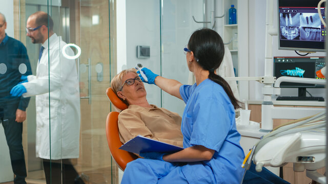 Nusre measuring old woman temperature before dental examination waiting for doctor. Dentist assistant interrogating senior woman and taking notes on clipboard sitting on stomatological chair