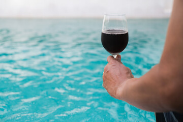Man hand holding glass of red wine sitting near the pool. Travel vacation. Summer holidays. 