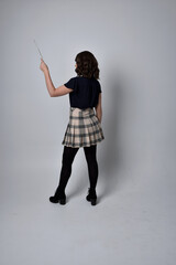 full length portrait of pretty brunette woman wearing tartan skirt and boots with long black cloak.. Standing pose holding a magical wand and casting a spell against a studio background.