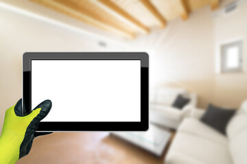 Hand with work glove holding a blank digital tablet with a blurred living room in the background.