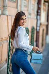 Outdoor waist up portrait of young beautiful girl posing in street. Model wearing white shirt, holding blue bag. City lifestyle. Female fashion concept.