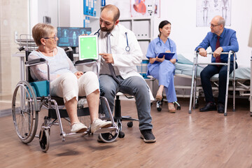 Handicapped senior woman sitting in wheelchair discussing with doctor holding tablet pc with green screen. Medical practitioner using device with chroma key during coonsultation of invalid senior