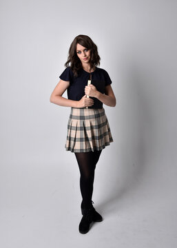 full length portrait of pretty brunette woman wearing tartan skirt and boots.  Standing pose holding books against a  studio background.