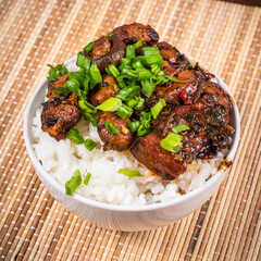 Small bowl with spicy stewed pork with mushrooms in oriental style with spices, hot peppers and boiled rice sprinkled with chopped green onions on a bamboo napkin and chopsticks - top view, close-up
