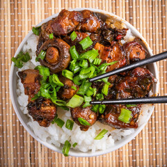 Small bowl with spicy stewed pork with mushrooms in oriental style with spices, hot peppers and boiled rice sprinkled with chopped green onions on a bamboo napkin and chopsticks - top view, close-up