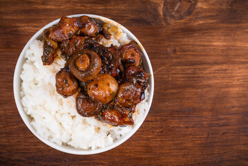 Small bowl with spicy stewed pork with mushrooms in oriental style with spices, hot peppers and boiled rice on a wooden table - top view, copy space, close-up
