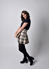 full length portrait of pretty brunette woman wearing tartan skirt and boots.  Standing pose holding books against a  studio background.