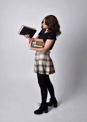 full length portrait of pretty brunette woman wearing tartan skirt and boots.  Standing pose holding books against a  studio background.