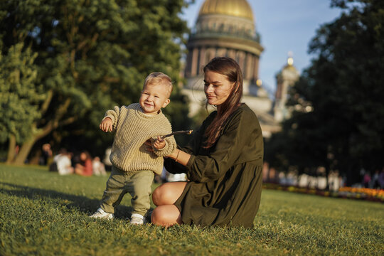 Young Caucasian lady, mother of little son sitting on grass in front of st isaac’s cathedral and helping boy stand holding him by hand. Image with selective focus