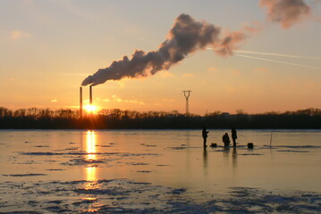 View of a frozen and snowy river at sunset  with a bridge in the background and white smoke from the chimneys.