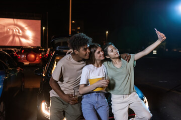 Cheerful diverse young friends smiling while taking a selfie together, standing in front of a big screen, ready to watch a movie in an open air cinema