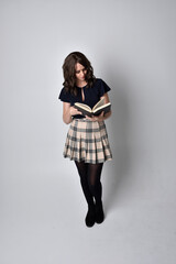 full length portrait of pretty brunette woman wearing tartan skirt and boots.  Standing pose holding books against a  studio background.