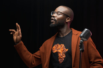 portrait of a dark-skinned handsome guy in brown jacket and black t-shirt stands with a microphone and emotionally sings in a studio on dark background
