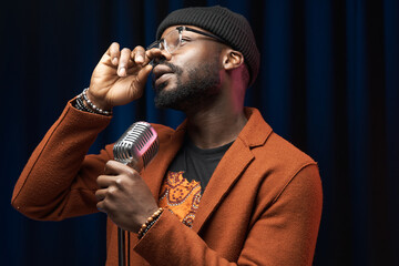 portrait of a dark-skinned handsome guy in brown jacket and black hat stands with a microphone and emotionally sings in a studio on dark background