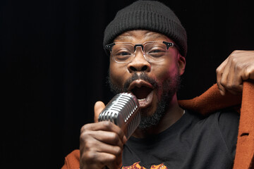 portrait of a dark-skinned handsome guy in brown jacket and black hat stands with a microphone and...