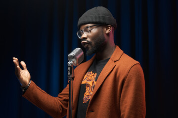 portrait of a dark-skinned handsome guy in brown jacket and black hat stands with a microphone and emotionally sings in a studio on dark background - 405083597