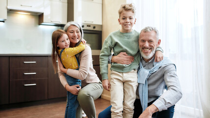 Cheerful family. Grandparent with grandchildren having fun and hugging each other in dining room or kitchen at home, posing for photo and smiling at camera