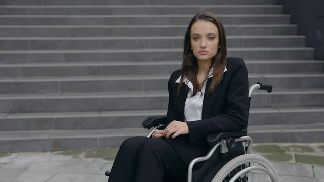 Crop view of handicapped beautiful businesswoman in suit sitting in wheelchair at street. Disabled young female person looking to camera outdoors. Concept of successful people.