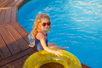 little girl in sunglasses and swimsuit, sitting by the pool in summer