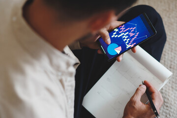 Rearview of a businessman holding smartphone with graphs and diagrams for marketing development. A male analyzing startup statistics, checking financial trade report presentation sitting at home.
