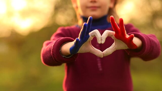 Love and happiness concept. Cute child forming heart gesture with hands outdoors on nature sunset bokeh background. Heart shape of kids hand painted in france flag colors, kids body language