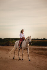 Cute happy young woman on horseback in summer beach by sea. Rider female drives her horse in nature on evening sunset light background. Concept of outdoor riding, sports and recreation. Copy space