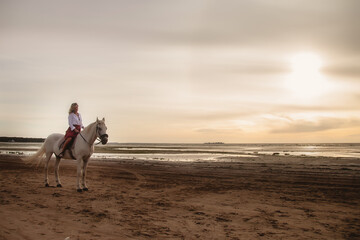 Cute happy young woman on horseback in summer beach by sea. Rider female drives her horse in nature on evening sunset light background. Concept of outdoor riding, sports and recreation. Copy space