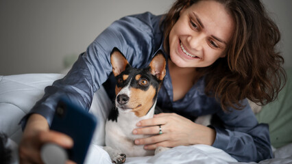 Beautiful young adult woman in pajamas doing selfie on smartphone with her dog bassengi in the morning in bed