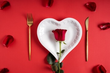 Romantic table setting for Valentines day. Love layout made with golden knife and fork, heart shaped plate and rose on red background. Minimal Valentines day or wedding dinner. Flat lay, top view.