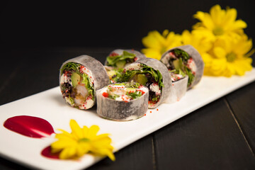 Side view of healthy Japanese rice free sushi roll with tempura shrimp wrapped in daikon radish. Yellow camomile flowers, red painting brush stroke decoration on white plate. Black Wooden background
