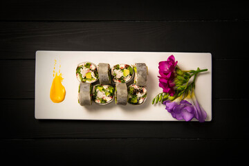 Top view of healthy Japanese rice free sushi roll with crab sticks wrapped in daikon radish. Colorful flowers, yellow painting brush stroke decoration on white rectangular plate. Black background
