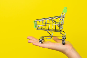 Empty shopping cart on female hand. Shopping theme. Small Trolley isolated on a bright yellow background. Consumer concept, Copy space.