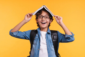Funny male pupil having fun with the book on his head ready for school isolated over yellow background