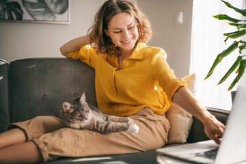 Casual woman in yellow shirt working on laptop with her cat on sofa, sitting together in modern room with window ang green plan