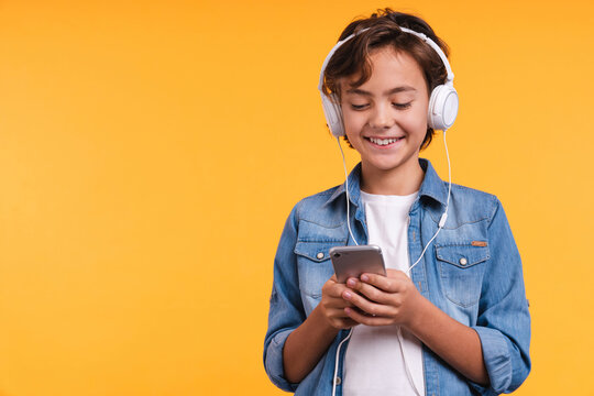 Cheerful young boy listening to the music in headphones and mobile phone isolated over yellow background