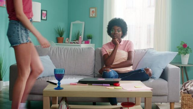 Young Afro-American woman in casual outfit is sitting on sofa, feeling sad, missing her friends and family, her roommate joins her and is trying to comfort and cheer, female friendship, Slow motion.