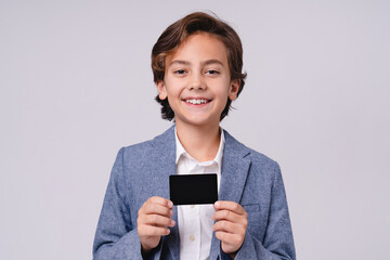 Handsome little 10s boy in formal attire presenting credit card isolated over grey background