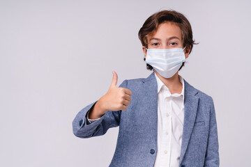 Smart young boy in medical mask against coronavirus showing thumb up isolated over grey background