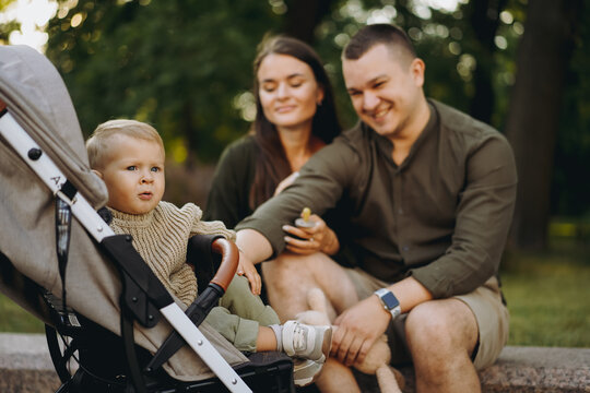 Baby boy sitting in a stroller, his young parents on background, sitting at ￼stone balustrade, looking at him with tenderness. Image with selective focus.