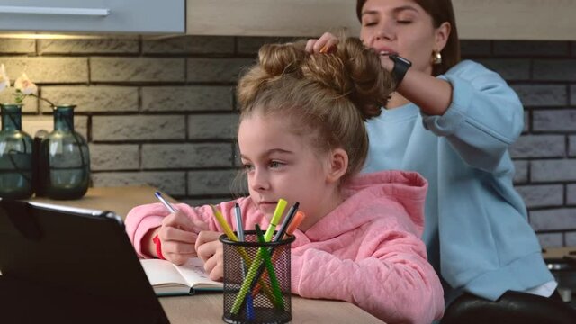 Careful mother is correcting her daughter's hair while the girl is drawing and using a tablet