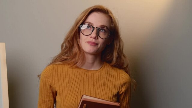 Freckled ginger woman is thinking a bout something while speaking on a gray wall holding a book and smile