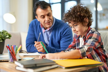 Father's help. Caring middle aged latin father helping, discussing homework with his son, school child while sitting together at the desk at home