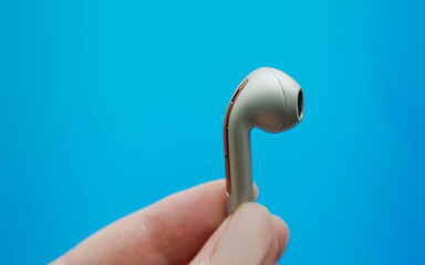 Close up of hand holding golden beige bluetooth earphones or wireless earbuds isolated on blue...