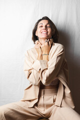 Gentle portrait of a beautiful young woman with big breasts in a beige suit and white lace bra sits on a chair on a gray white background