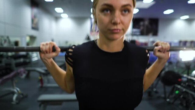 Fit blonde woman working out with barbell and looking at camera, equipment on blurred background. Camera tilt up and down female weightlifter in gym. Concept of sport